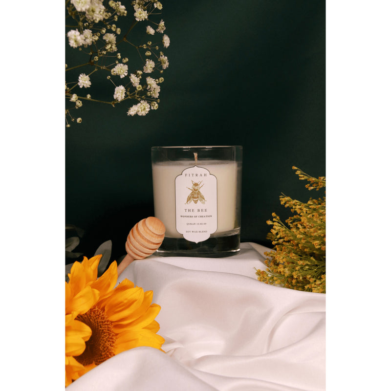 the bee candle from the wonders of creation collection on top of a white cloth with flowers and a honey dipper on the side.